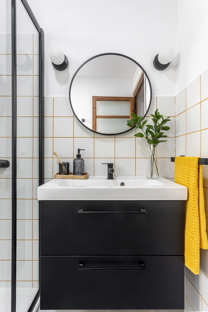 Detalle del baño con espejo circular, mueble negro y acento amarillo